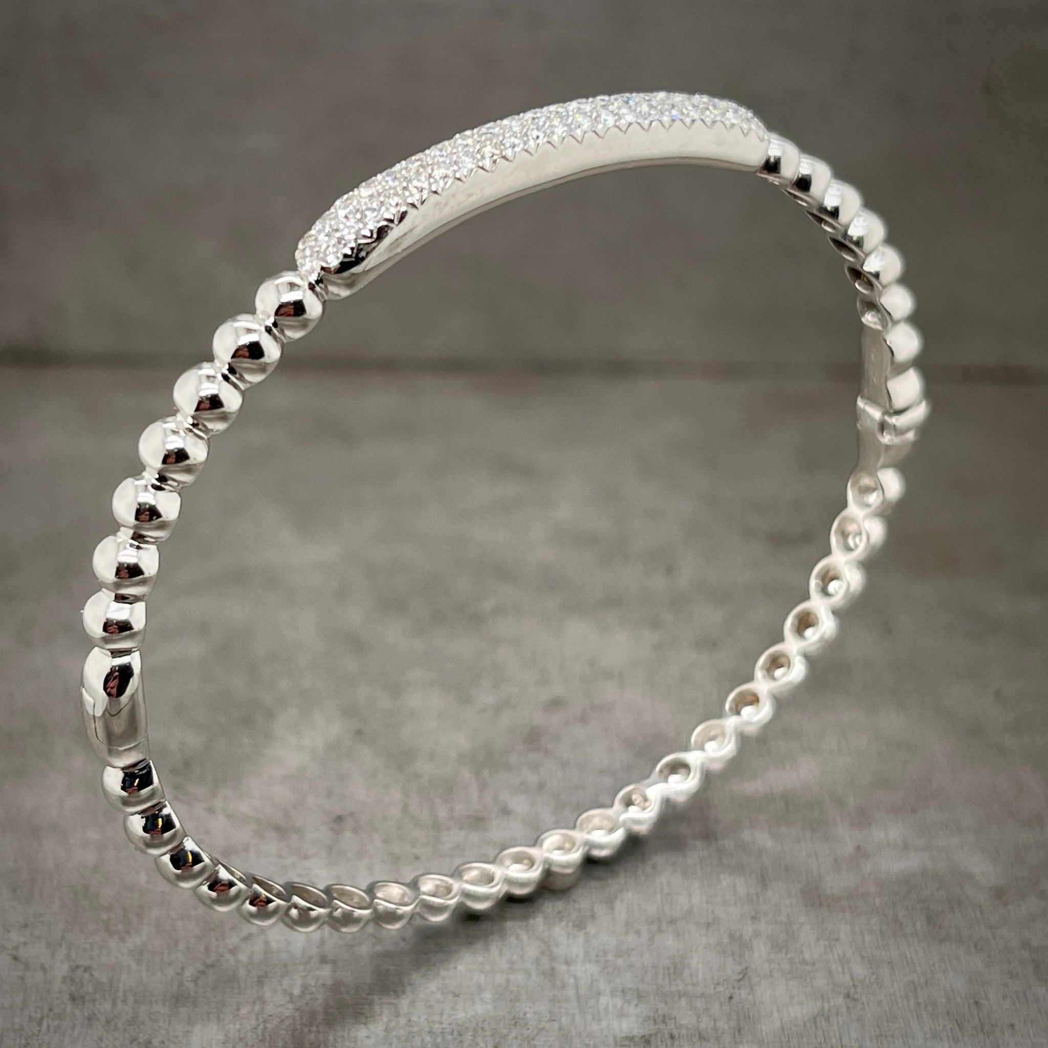 Full angled view of white gold diamond bangle standing up. Here you can see a bar that has multiple rows of pave set diamonds at the top of the bracelet, covering 1/5 of the circumference. The rest of the bangle is composed of balls of white gold.