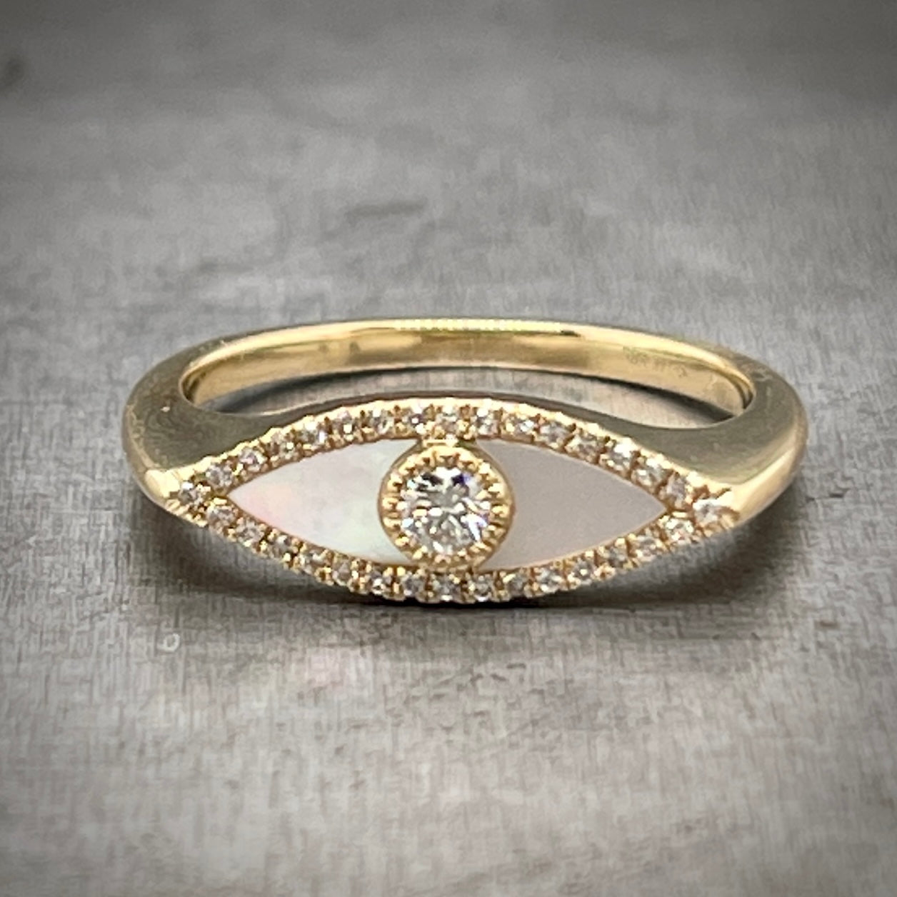 Full view of mother of pearl evil eye ring laying down on a gray background. The ring is made of yellow gold. The top of the ring features an eye ike shape made from a halo of round diamonds on the outside, mother of pearl creating the sclera of the eye and a round diamond at the center creating the pupil/iris.