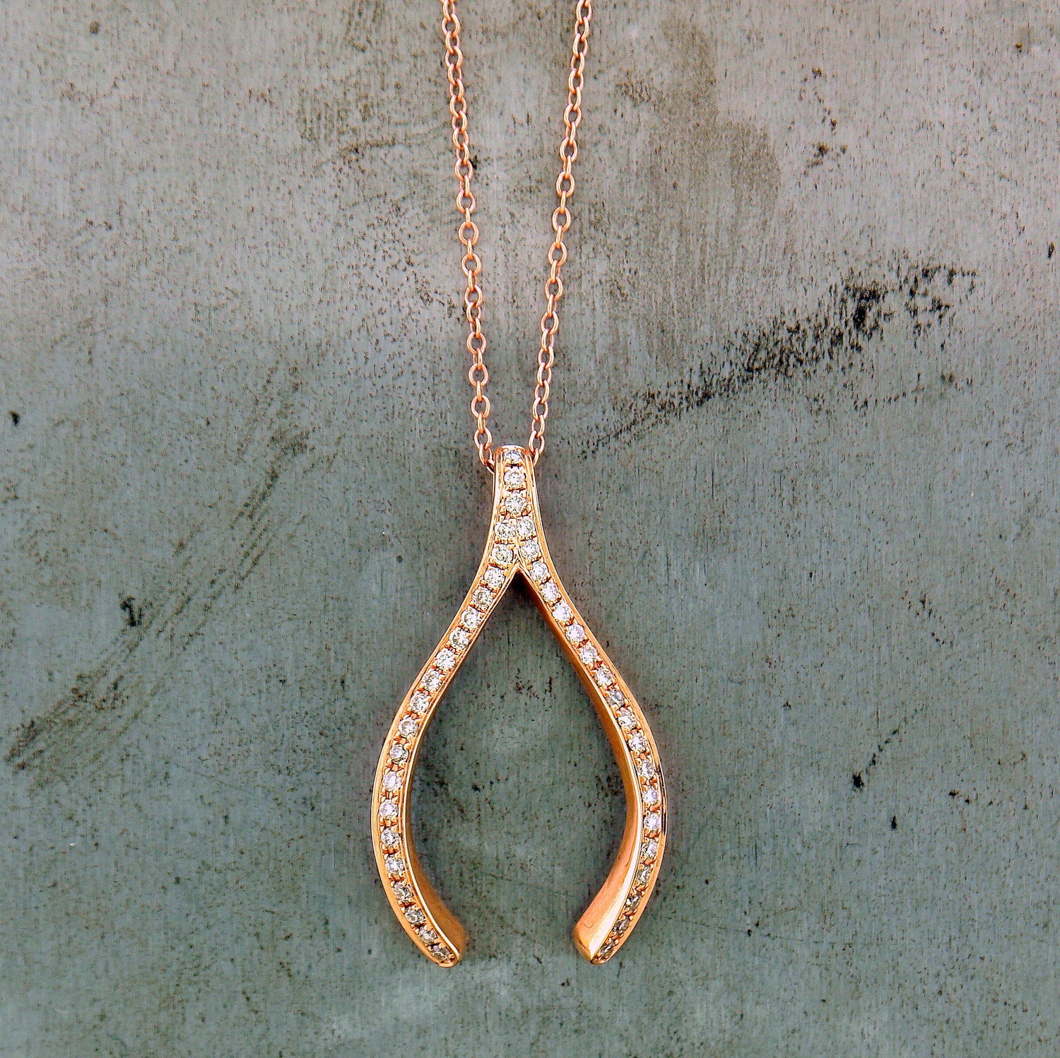 14 karat rose gold pendant in the shape of a wishbone. The tail end of the wishbone acts as the bail of the pendant. Covering the face of the wishbone are pave set round brilliant diamonds. There is only one row of diamonds on the face of the wishbone. The pendant lays on a 14 karat rose gold curb chain. This pendant lays on a gray background.