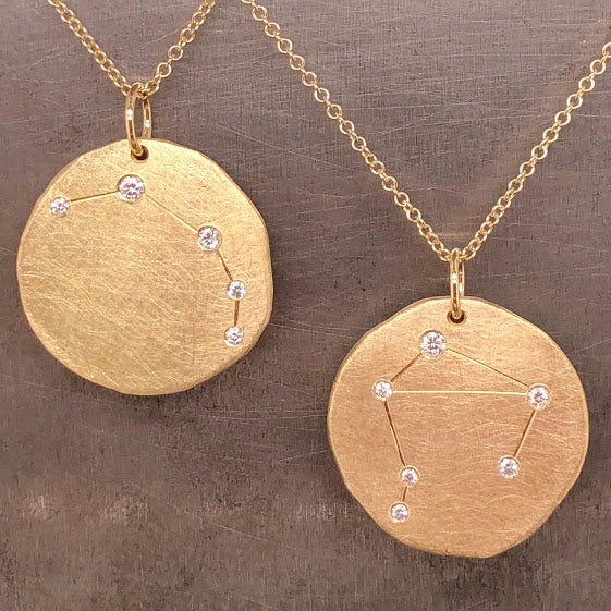 Close up view of two zodiac pendants. The pendants are organic circles that feature lifework for the zodiac and round diamonds set on each angle of the zodiac. The left pendant features Aries with five diamonds. The right pendant features Libra with six diamonds.