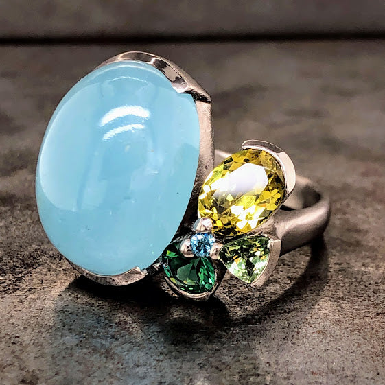 Angled view of Aquamarine 18 karat white gold ring laying down. To the left is an oval aquamarine cabochon partial bezel set on its Northern and Southern girdles. To the left are four sapphires. There is one oval cut light yellow-green sapphire partial bezel set. Next to that sapphire is a trillion cut sage green sapphire partial bezel set. Next to the trillion is a round deep green sapphire. In the middle of these three sapphires is one small round blue sapphire.