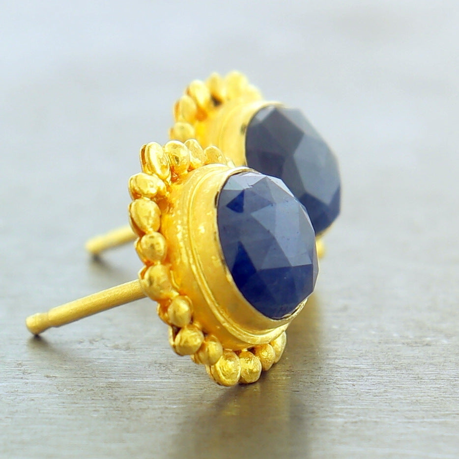 Side view of 22 karat yellow gold blue corundum post earrings. The blue corundum is round cut and bezel set in gold. Surrounding the bezel are irregular shaped bubbles made from gold.