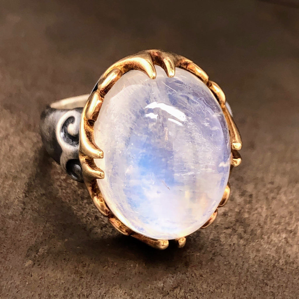 Angled view of 18 karat yellow gold moonstone ring. This ring features an oval cut cabochon moonstone. This moonstone is claw prong set in 18 karat yellow gold. The setting looks very ancient with swirling motifs etched into it and the claw prongs in groups of two. The moonstone is set with four groups of claw prongs. The shank of the ring is made from oxidized sterling silver and showcases a swirling motif in its shoulder.