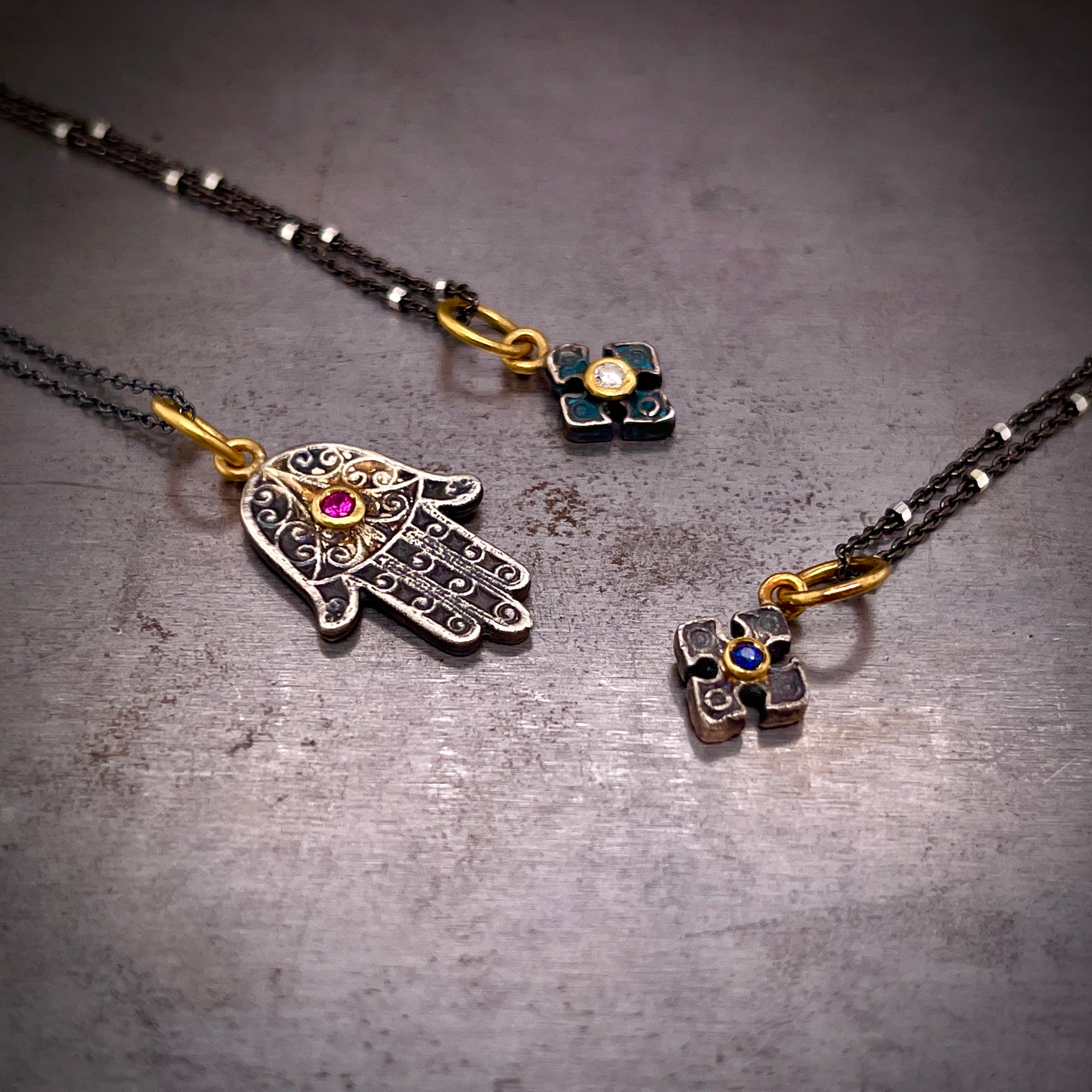 Close up view of three oxidized sterling silver gemstone pendants. Two pedants come in angled from the left and one from the right. The top left features an antique cross-like pendant made from oxidized silver with a blue coloring. This pendant features a round brilliant diamond bezel set in yellow gold. Below is a hams hand with a bezel set ruby. To the left is another cross with a blue sapphire set in yellow gold.