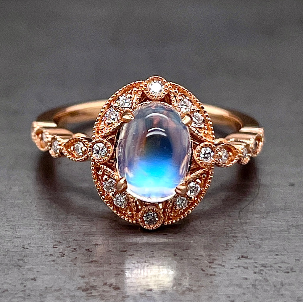 Frontal view of ring. An oval cabochon moonstone is four prong set in 14 karat rose gold in the center. There are 12 round brilliant diamonds that surround the moonstone; one at each corner and then groups of two in-between each corner diamond. The shoulders of the shank feature round brilliant diamonds that are encompassed by metal to make the shape of a marquise, then a circle, then a marquise, and then a circle again. Milgraine detailing is features on all the edges.