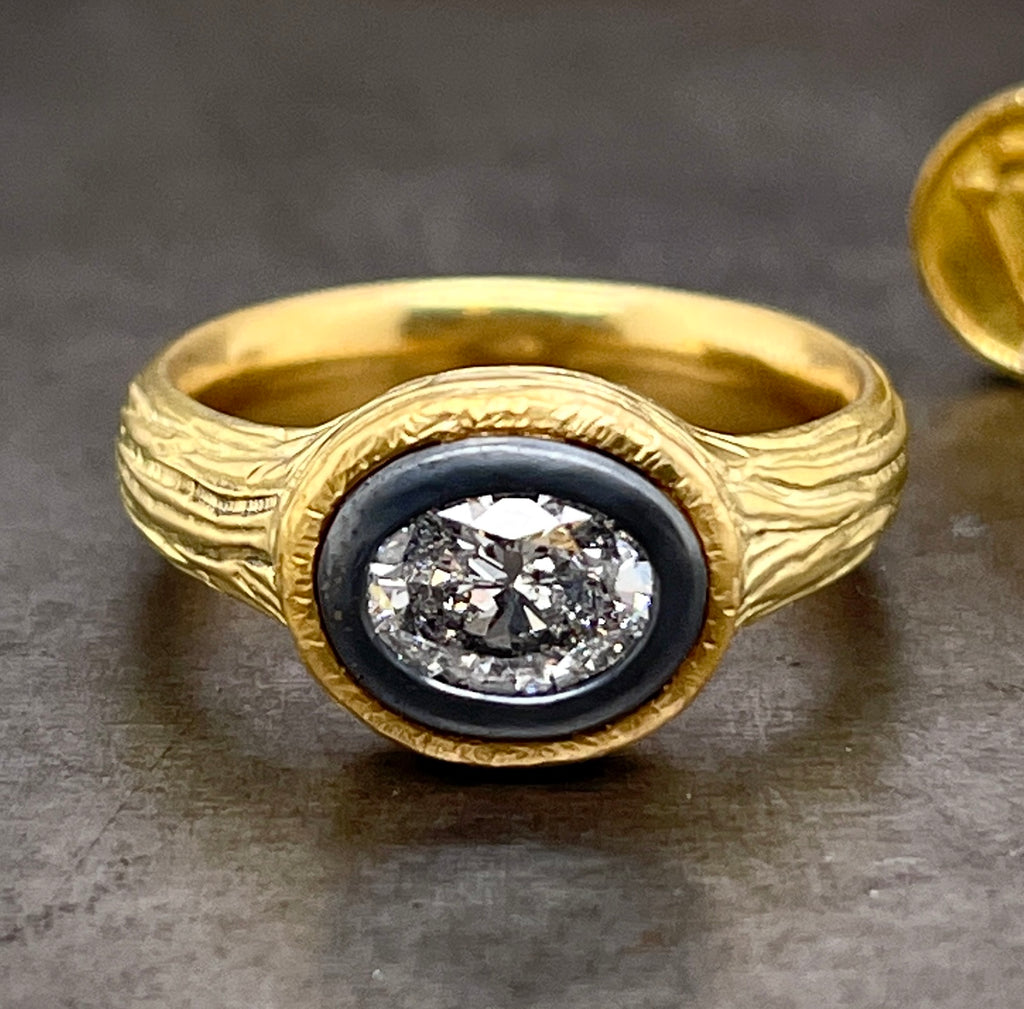 Frontal view of salt and pepper diamond ring. An oval cut diamond with white and black inclusions is bezel set in oxidized sterling silver. Surrounding the silver is 18 karat yellow gold with bark texture.