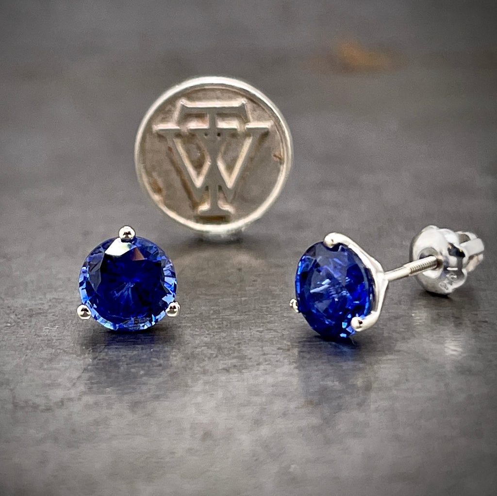 Frontal and side view of AAA Quality Sapphire studs. On the left you see a round blue sapphire (a deep blue) three prong set in white gold. On the right you see the side view of the studs where you can see the screw on post. In the back there is a "WT" coin standing up.