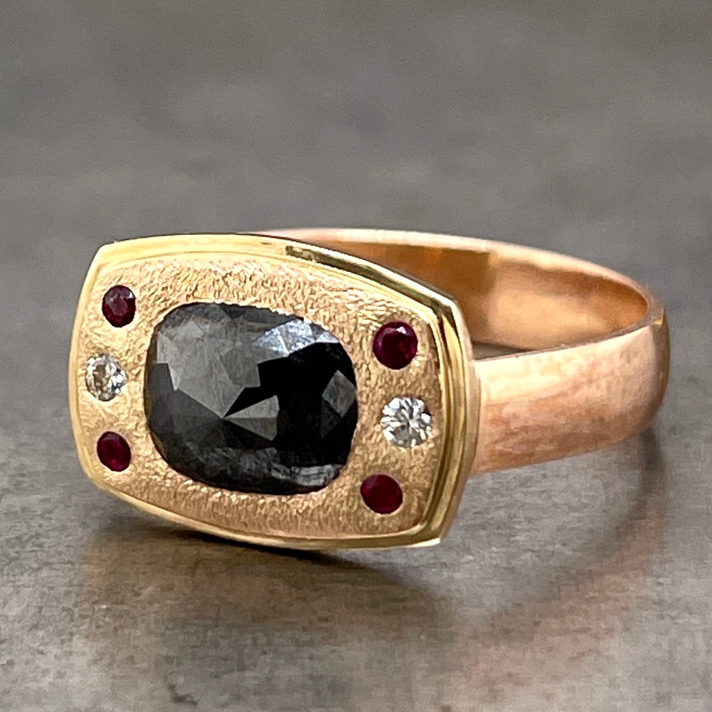 Angled view of Black Diamond and Ruby Ring laying down. The face of this ring features a rounded rectangle of 14 karat yellow gold. In the center is a rose cut rectangle black diamond flush set. To the left and right of the black diamond are three stones; a round ruby, round brilliant diamond and another round ruby. This head lays on a 14 karat rose gold band.