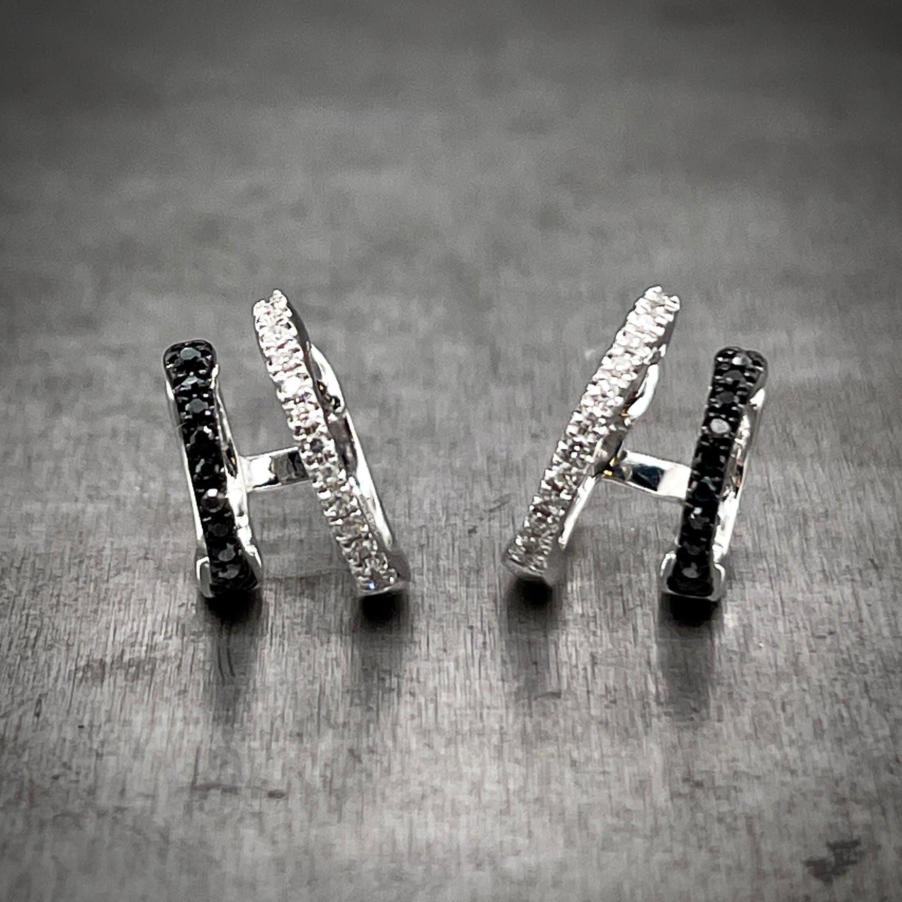 Full view of black diamond hoop huggies. The inner most part of the earring features a larger hoop huggies that has round brilliant diamonds set in it. The outer hoop is slightly smaller and features black diamonds. The hoops are attached together in the back by a single bar so when worn, the earrings give the illusion of a double piercing.