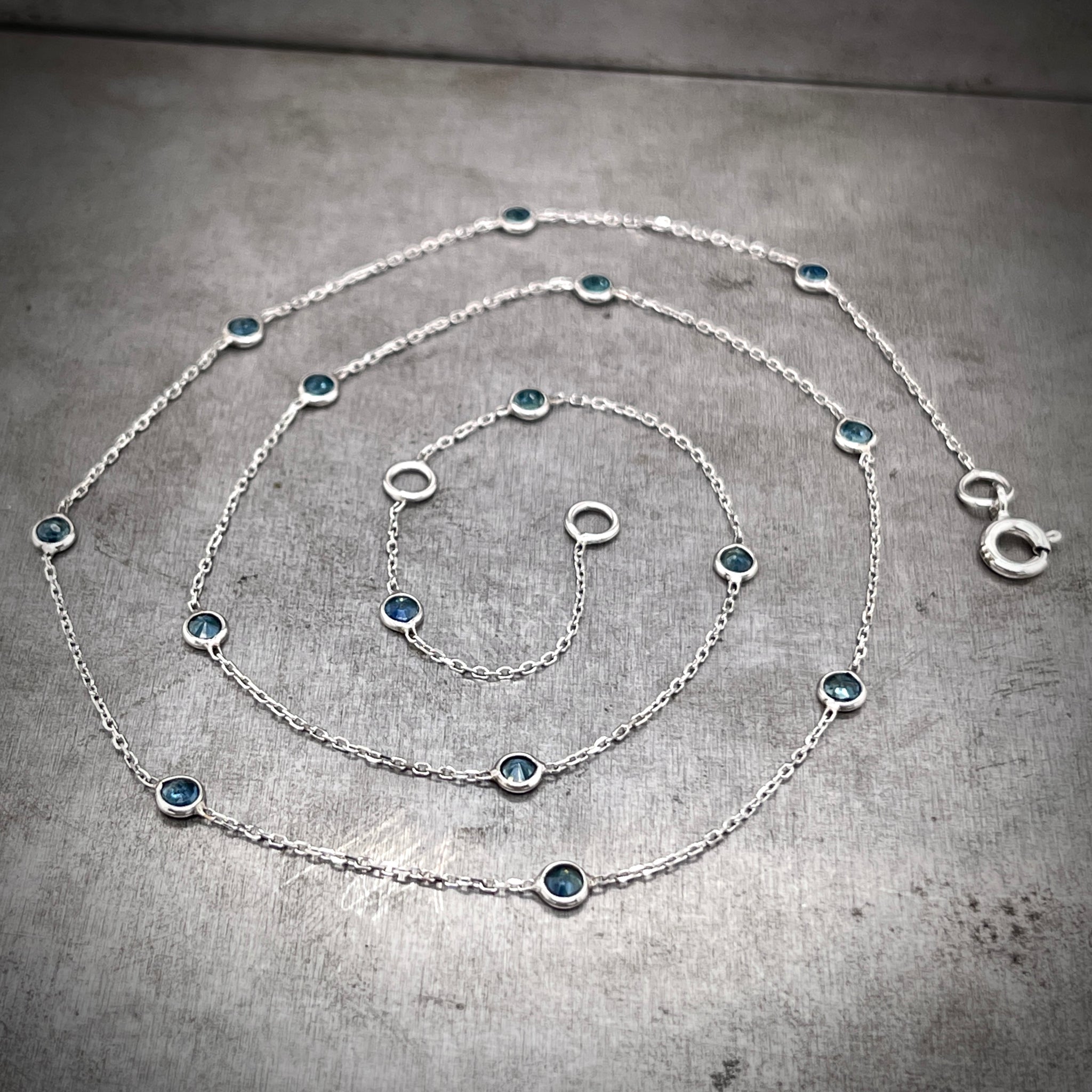 Full view of White Gold and Sapphire Necklace laying in a spiral on a gray steel background. About every inch this chain features a small rose cut blue sapphire that is bezel set in white gold. This necklace features 15 sapphires.