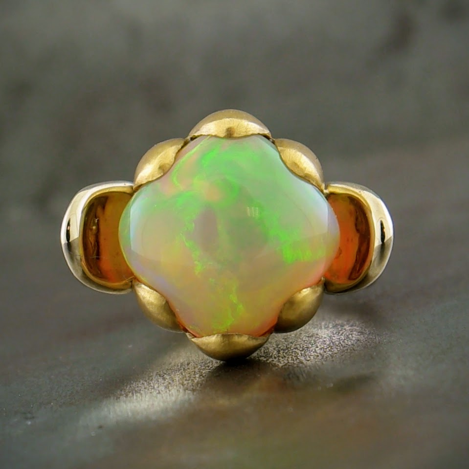 Full view of top of ring. A four leaf clover like shape of an Ethiopian opal is in the center. This opal is milky white with streams of neon green, neon yellow, orange and violet throughout it. This opal is six prong set. These prongs are grouped in three's, one set on the top and the other on the bottom. These prongs are in the shape of curving flower petals, curving around the opal. To the left and right of the opal is a larger leaf like setting made from 18 karat white gold. 