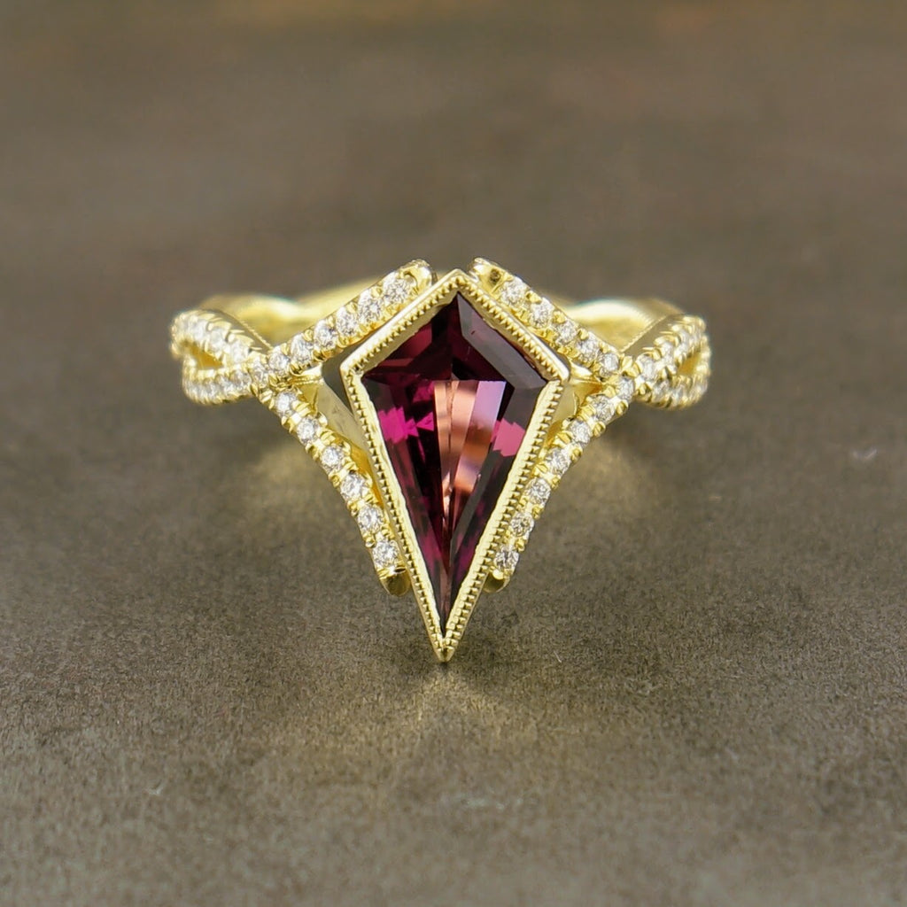 Frontal view of sapphire and diamond yellow gold ring. A kite like diamond deep red sapphire is set in the center of the ring in a bezel with milgraine detailing on the edges. The ring features a woven shank that spreads around the kite sapphire like a "V" on either side. The shank features a row of round brilliant diamond prong set.