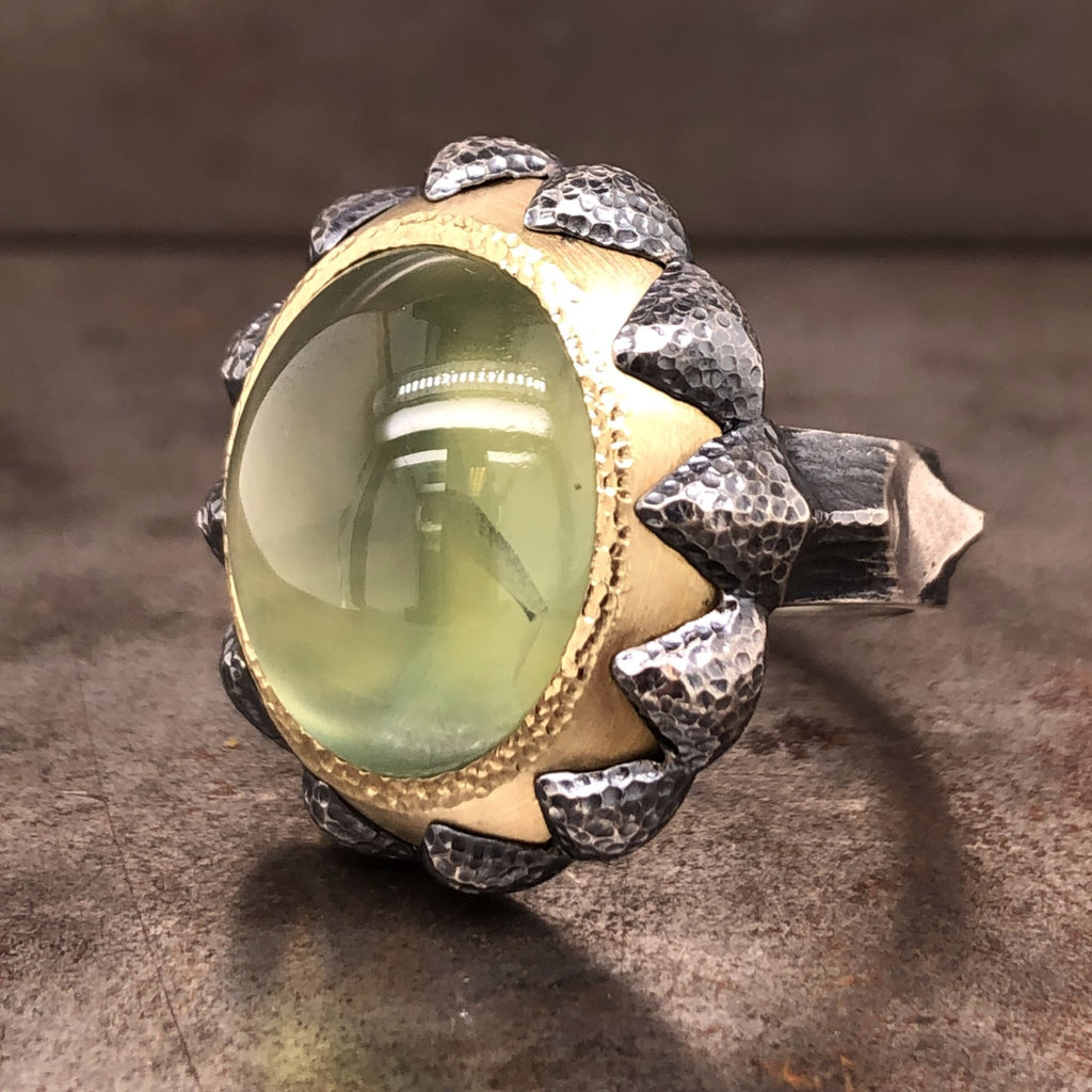 Angled side view of ring. Features one oval cabochon light green prehnite set in 18 karat yellow gold. There are claws made from oxidized sterling silver that reach over the yellow gold. The silver features a hammered texture.
