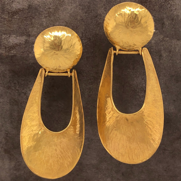 Front view of 18 karat yellow gold oyster shell earrings. These dangle earrings feature a circle disk of gold as the post. Hanging from a hinge off the bottom of the disk is an oblong horseshoe shape. The gold all features an oyster shell like texture from hammering the metal.