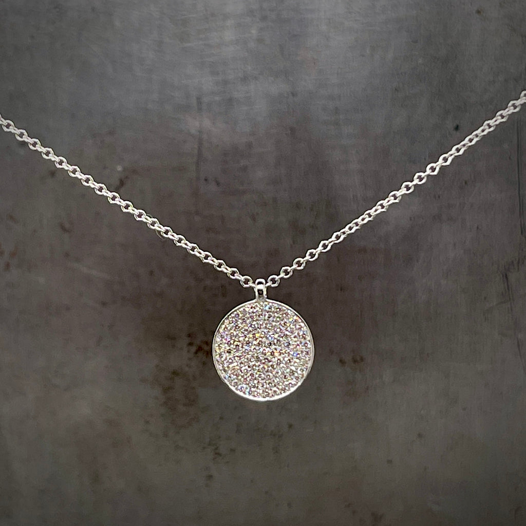 Close up view of Diamond Disc Pendant hanging with a gray background. The pendant is a circle that has diamonds covering the surface creating a sparkle disc.