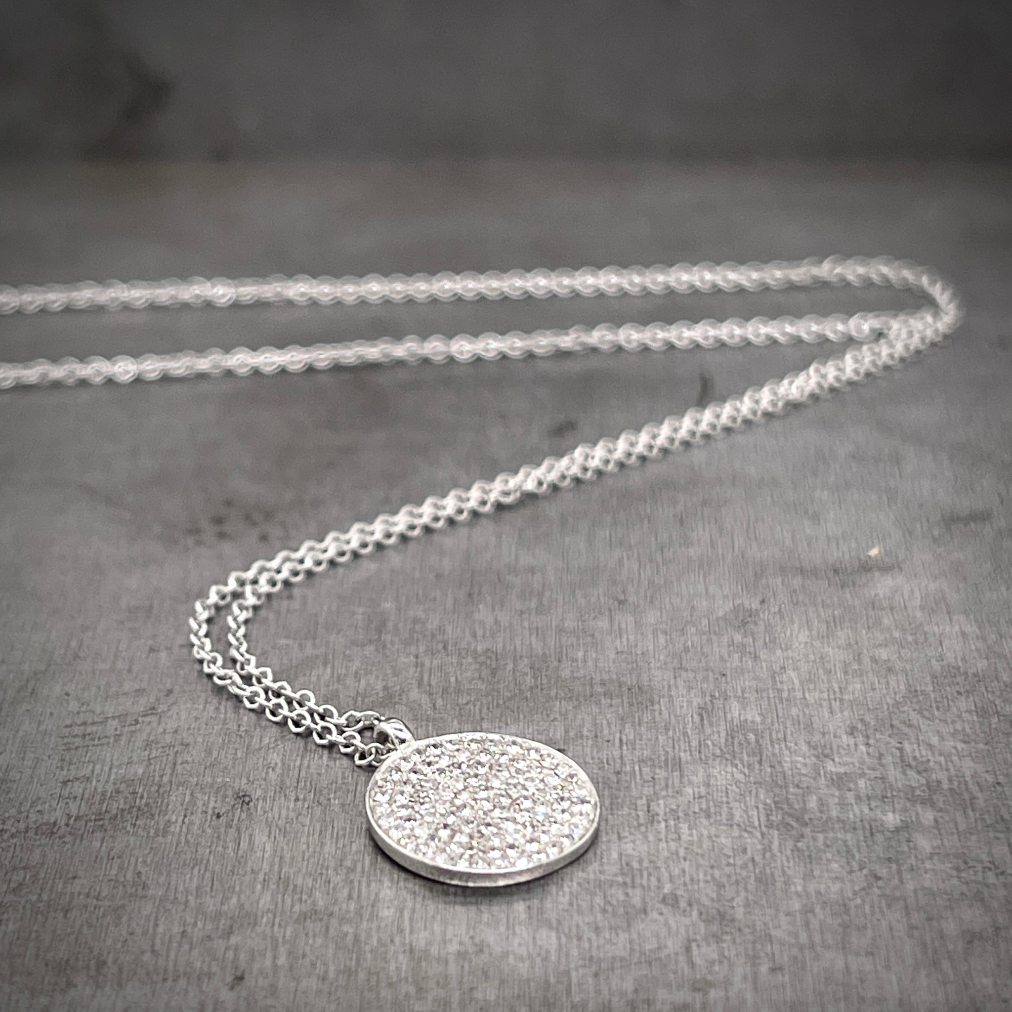 Full view of Diamond Disc Pendant laying on gray background with its chain twisting in the background.
