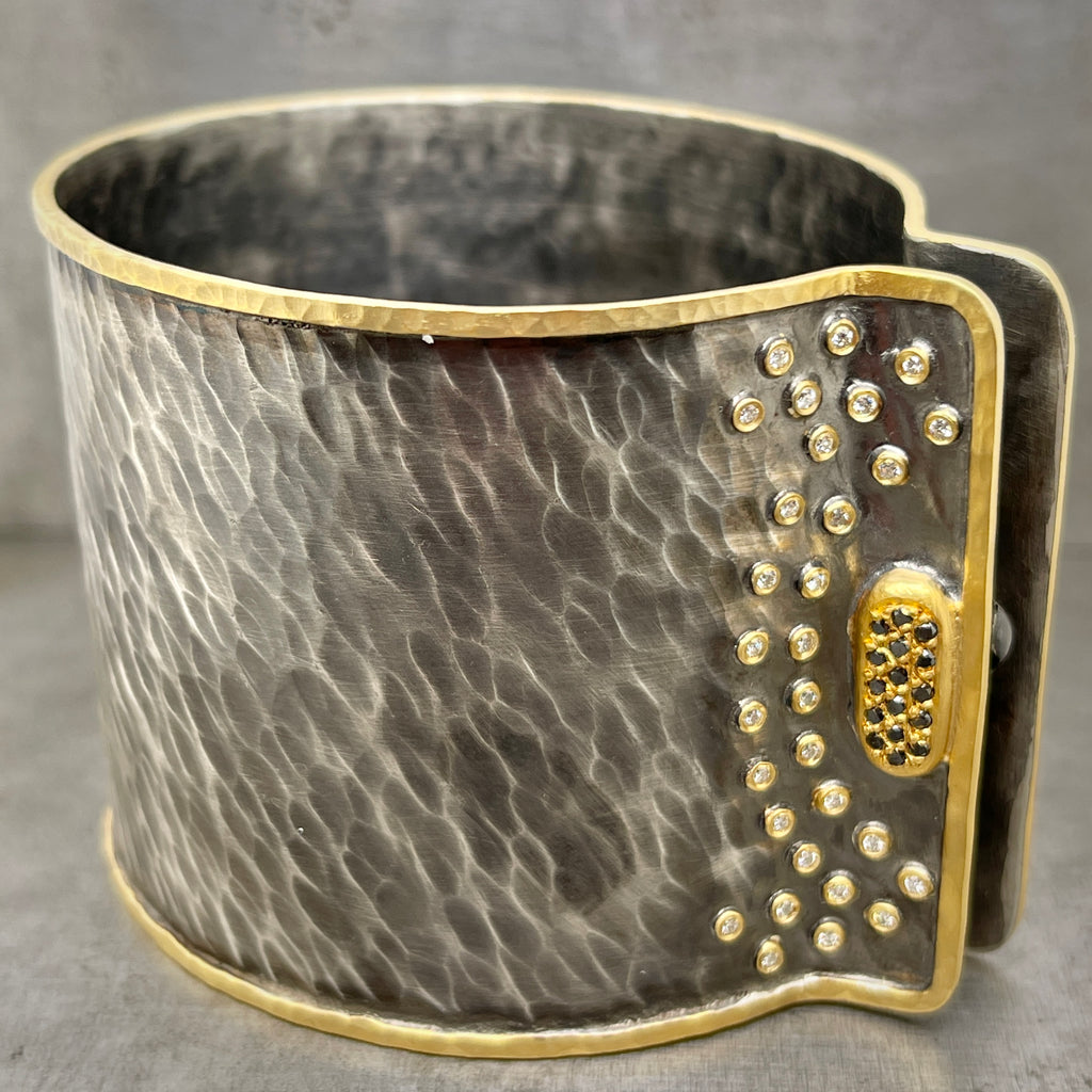 Full angled view of ancient oxidized cuff. This cuff features 30 diamonds that are bezel set in 22 karat yellow gold sporadically around the clasp of the cuff. This cuff is 2 inches tall and is made from oxidized sterling silver with a hammered finish and a border of 22 karat yellow gold. The clasp features 16 round black diamonds all set in 22 karat yellow gold.
