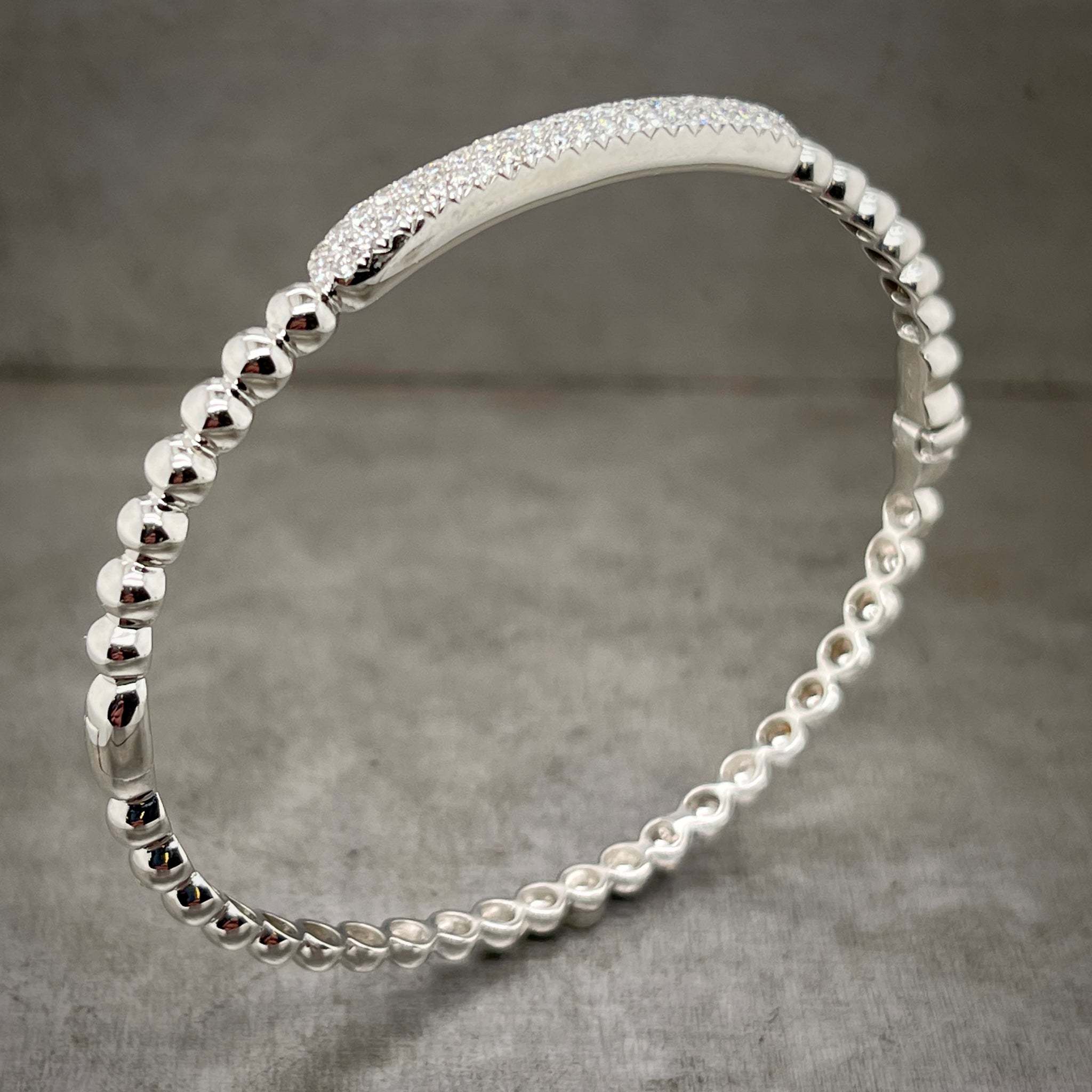Full angled view of white gold diamond bangle standing up. Here you can see a bar that has multiple rows of pave set diamonds at the top of the bracelet, covering 1/5 of the circumference. The rest of the bangle is composed of balls of white gold. 
