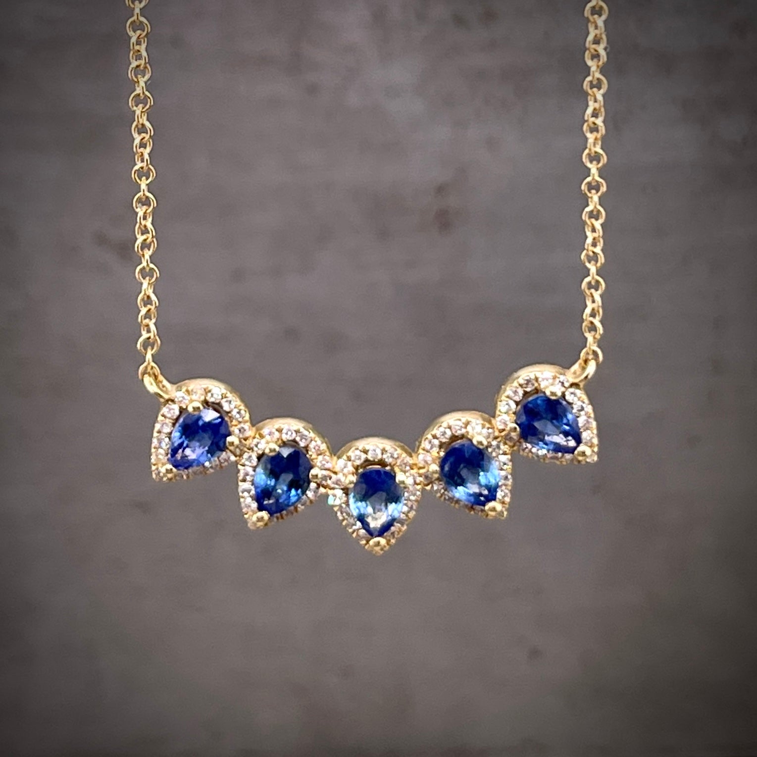 Close up view of pendant on pear and sapphire diamond necklace. Features 5 pear cut blue sapphires that create an upward curved line with their tips pointing south. Surrounding each sapphire is a halo of small round brilliant diamonds.