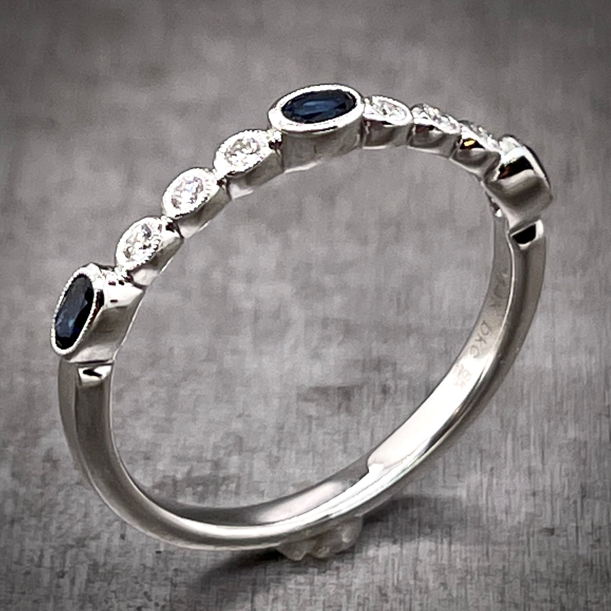 Angled aerial view of sapphire and diamond band. The three blue oval sapphires are spaced evenly across the top of the shank with three round diamonds in-between each sapphire. The stones only go around half of the shank. The bezels of each stone feature a beading detail on top of them.