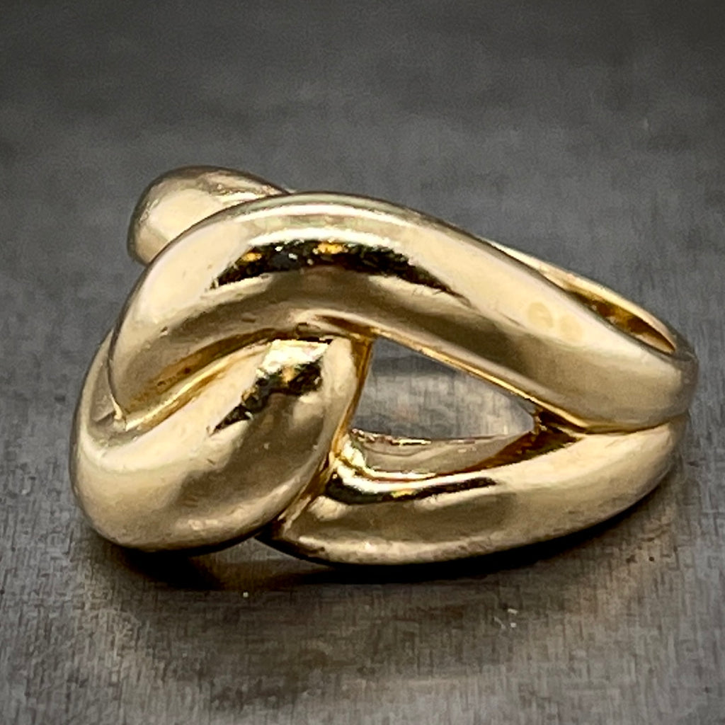 Angled side view of Knot Ring laying down on gray background. Looks like an endless knot with two loops that intertwine at the top of the ring and smooth out into one band on the bottom.