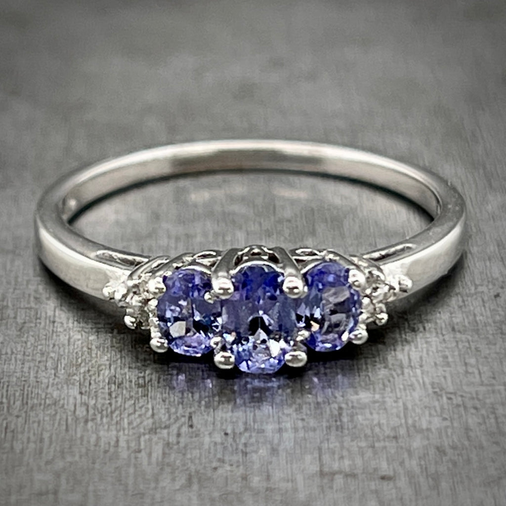 Full view of White Gold Tanzanite Ring laying on a gray background. There are three oval tanzanite set at the top of the ring, the center being the largest and the two flanking it being slightly smaller. Two single cut diamonds flank either side of the tanzanites.