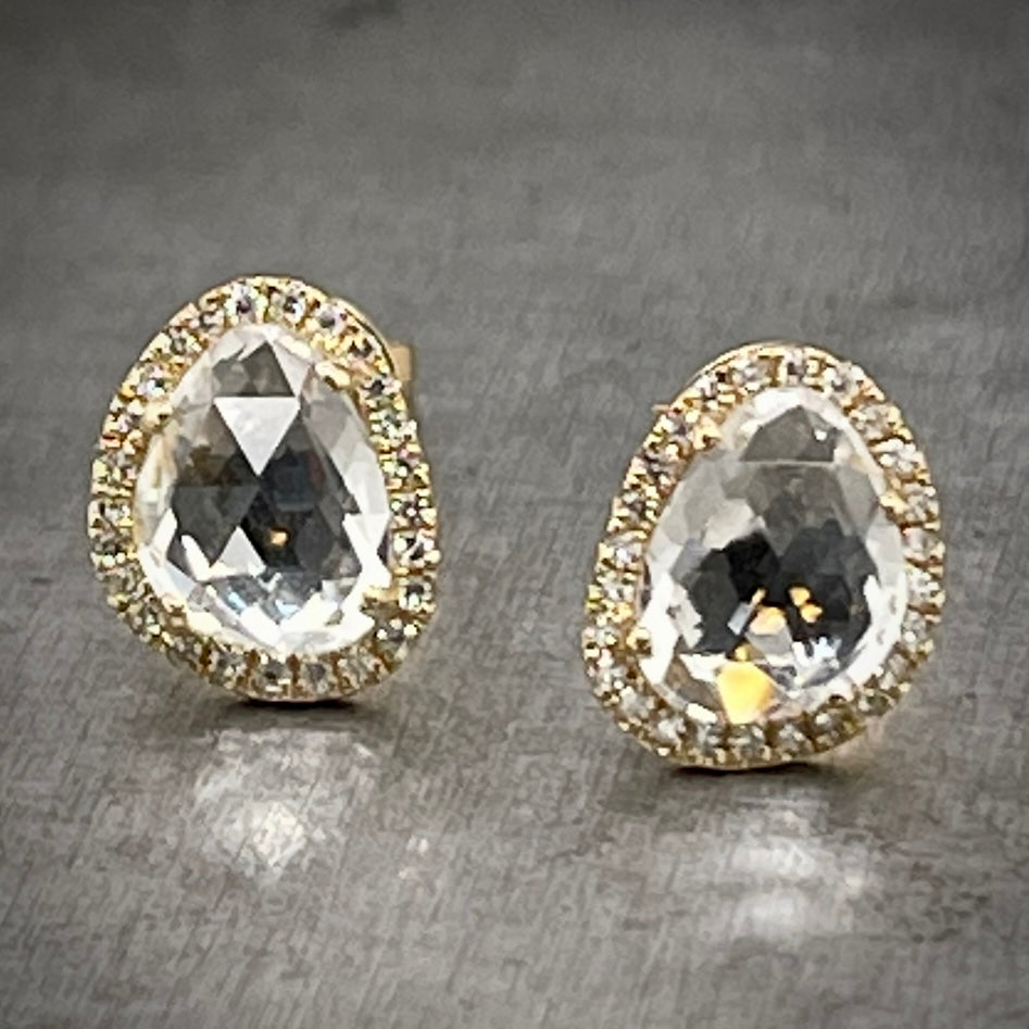 Full close up view of white topaz studs. These studs are made of yellow gold and each feature one oblong ovular shaped white topaz. This topaz is 4 prong set and features a halo of round diamonds surrounding it. These earrings lay on a gray background.