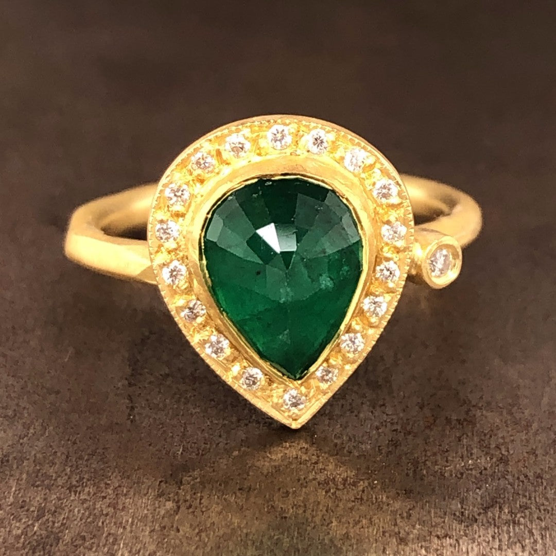 A pear rose cut emerald is featured in the center of the ring, with the tip pointing south. The emerald is bezel set with a rim of gold surrounding it. The rim features a halo of round brilliant diamonds that surround the emerald. To the right of the emerald and diamond head is a single round brilliant diamonds bezel set. Lays on a gold band.