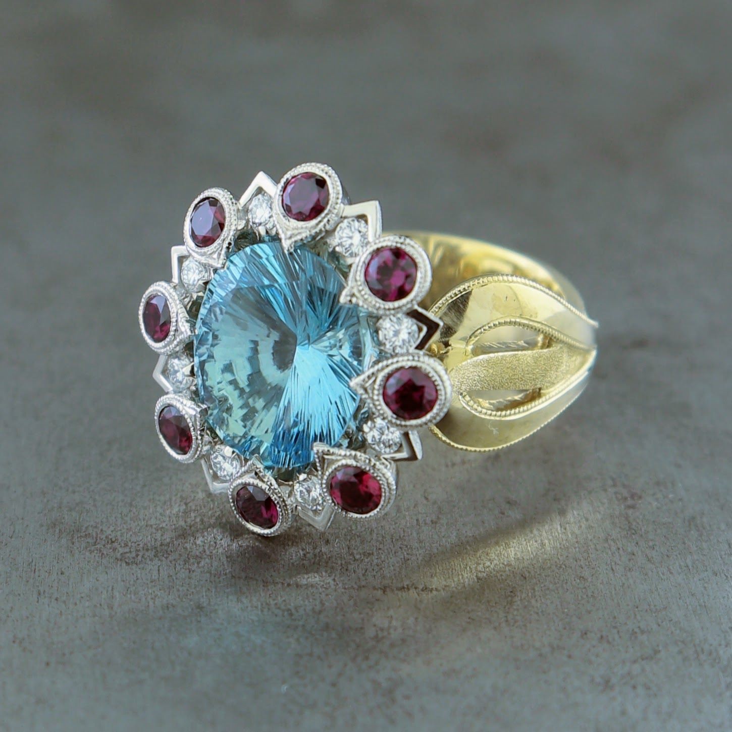Angled side view of firework aquamarine ring. Features a round baby blue sapphire set in the middle. Surrounding the sapphire is  halo of small round brilliant diamonds and then bigger round rubies. The mounting features a curved and the triangular shape to it that alternates from ruby to diamond the head is white gold. The shank is thick, curved and gold.