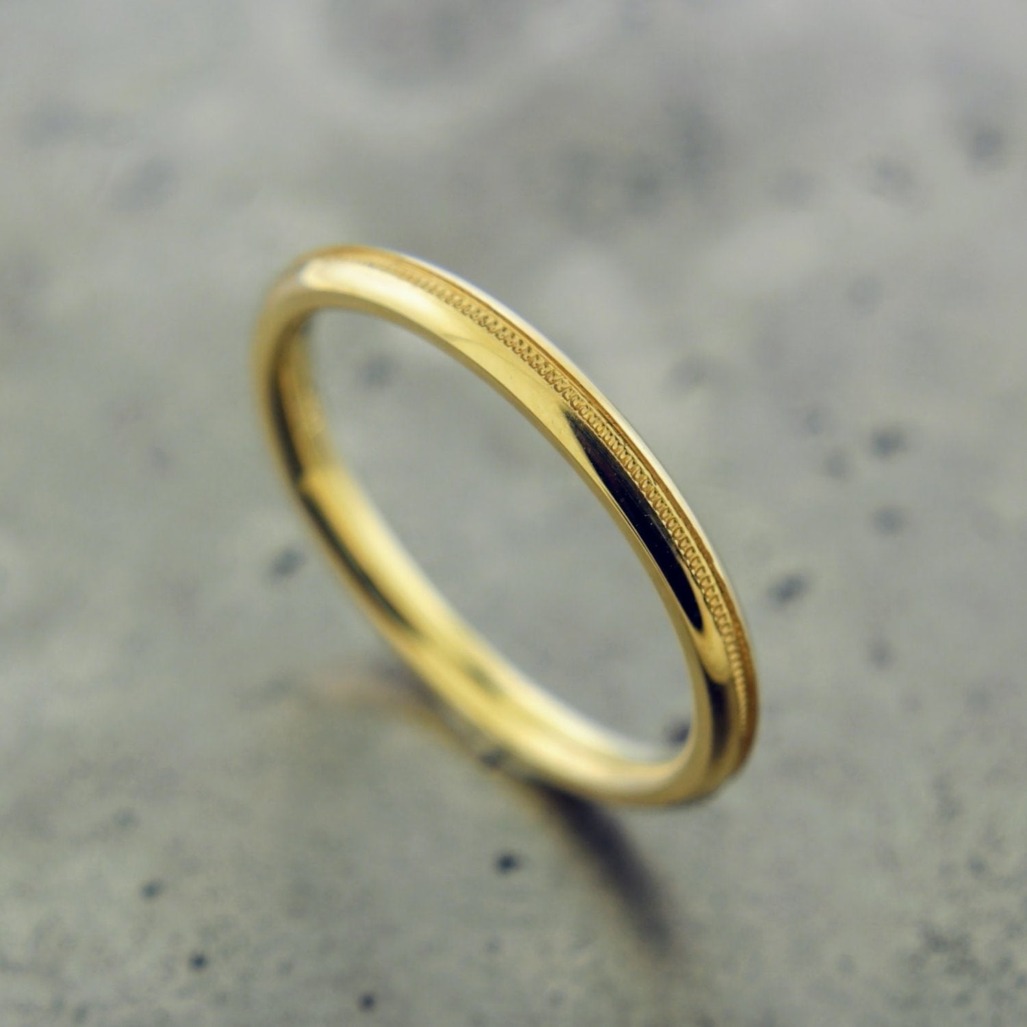 14 karat yellow gold ring. This ring features milgraine beading in the center of the band that circles the entirety of the ring. This ring features an angled knives edge to the ring, where it is angled about 45 degrees on the outer edges and showcases the milgraine detailing in the center of the knives edge. The ring features a high polished finish. This ring lays on a gray background.  
