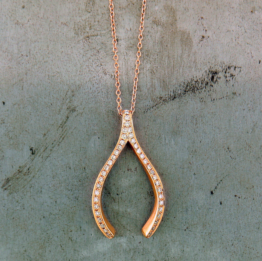 14 karat rose gold pendant in the shape of a wishbone. The tail end of the wishbone acts as the bail of the pendant. Covering the face of the wishbone are pave set round brilliant diamonds. There is only one row of diamonds on the face of the wishbone. The pendant lays on a 14 karat rose gold curb chain. This pendant lays on a gray background.