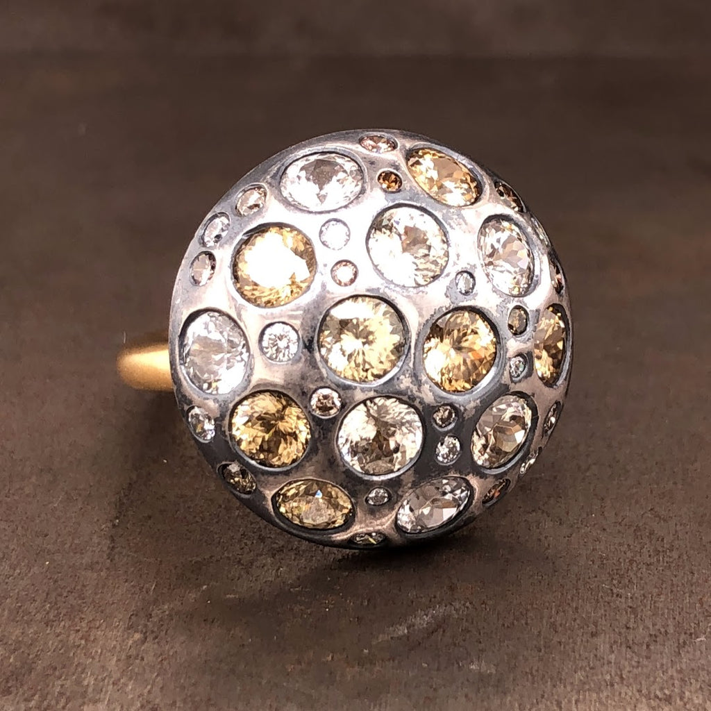 18k yellow gold and sterling silver shield ring with diamond and natural zircon. This ring features round brilliant diamonds and round zircon's flush set into a hemisphere of oxidized sterling silver. These stones have champagne coloring. This hemisphere lays on the shoulder of the ring; the shank of the ring is made from 18 karat yellow gold.