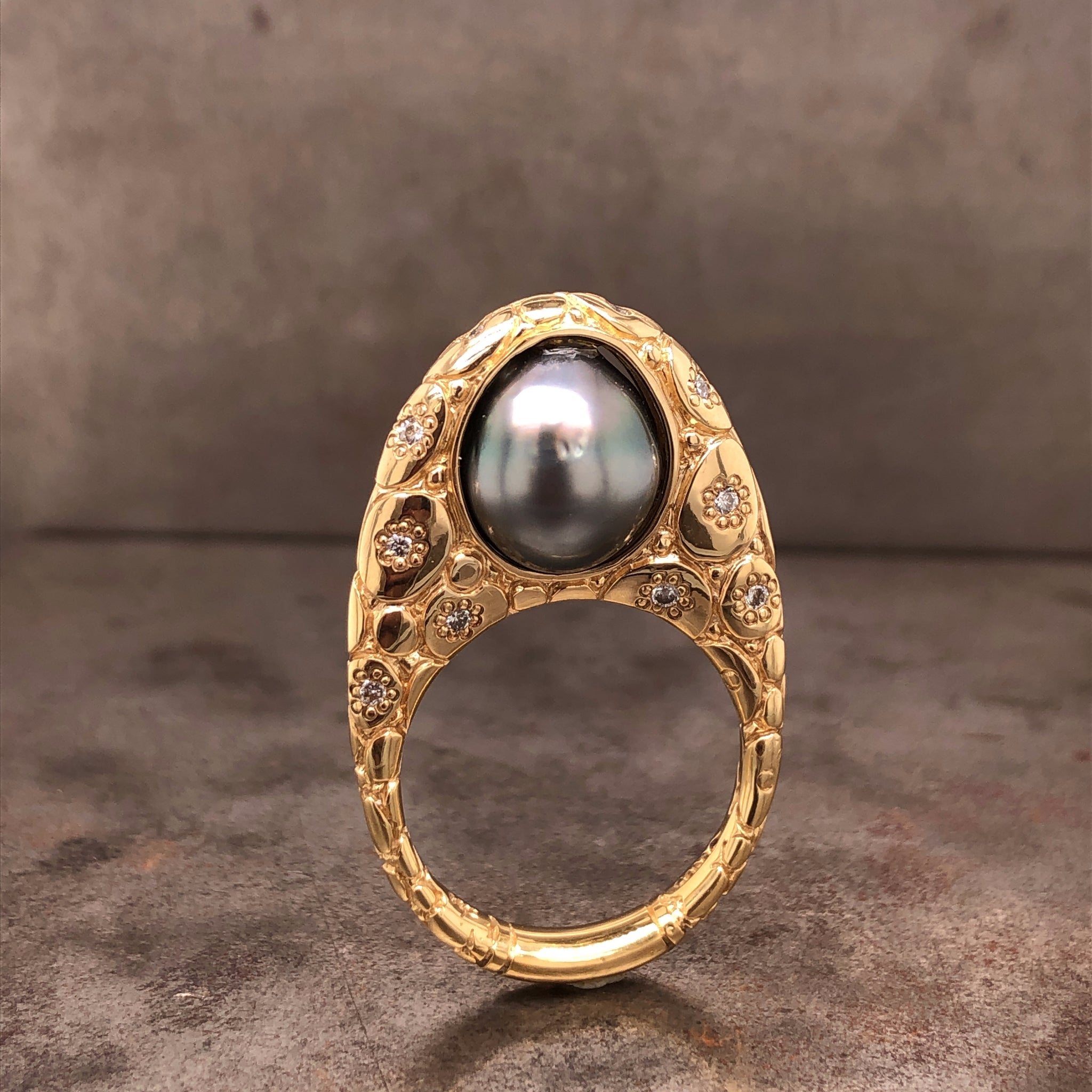 Full view of ring standing up. Ring profile looks like an egg, the opening for the ring is positioned near the bottom of the egg. Set in the center of the ring is a black Tahitian Pearl. This pearl is encompassed with gold. The rest of the egg mounting features carved portions resembling a cobblestone walkway and round brilliant diamonds scattered around the mounting.
