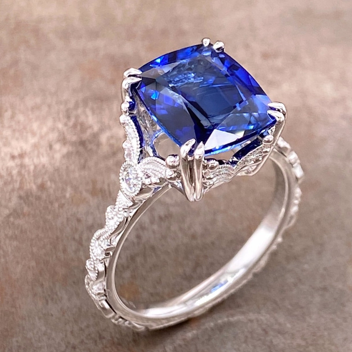 Aerial view of sapphire and 14 karat white gold ring. The ring features an emerald cut blue sapphire that is claw prong set with two claws at every corner. The shank features an Art Deco design with migraine on every border and a round diamond set on the shoulder of the shank.