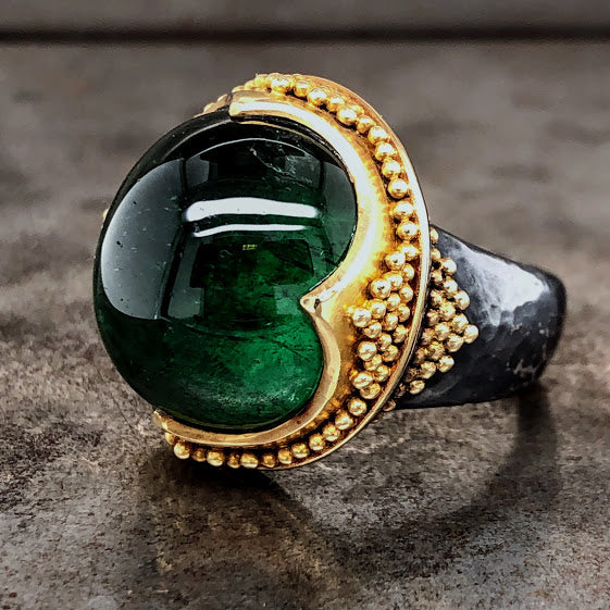 Side profile of green tourmaline ring. The tourmaline is a round cabochon cut and a deep green color. It is bezel set in yellow gold with curved antique looking setting that comes to a point at four corners. Within the bezel is beaded milgraine detailing. The shank is made from oxidized sterling silver and is hammered. There is gold milgraine detailing on the shoulder of the shank and makes a triangular form.