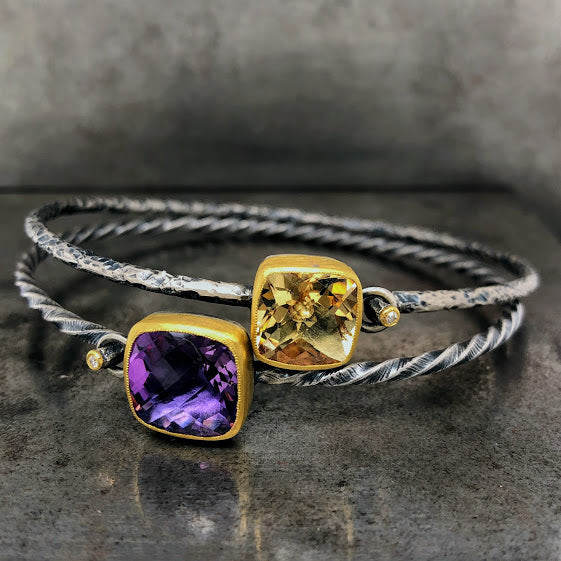 Full view of two bangles. The top bangle features an oxidized sterling silver cuff that looks hammered. The center of the cuff features a cushion cut citrine bezel set in 24K gold. On the tension part of the cuff there is a tiny round diamond set in gold. The cuff below if features an oxidized silver twisted cuff with a cushion cuff amethyst. It also features a diamond set in its tension/clasp.
