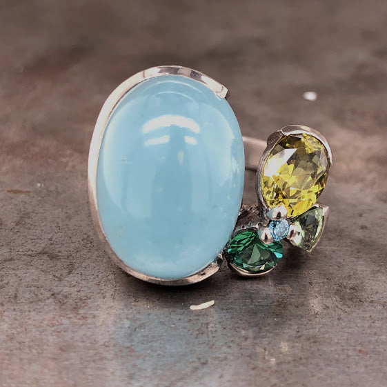 Frontal view of aquamarine 18 karat white gold ring. To the left you see the large cabochon aquamarine partial bezel set on three sides except for its Eastern side. To the left is the closer of four sapphires with the small blue sapphire set in the middle of them.