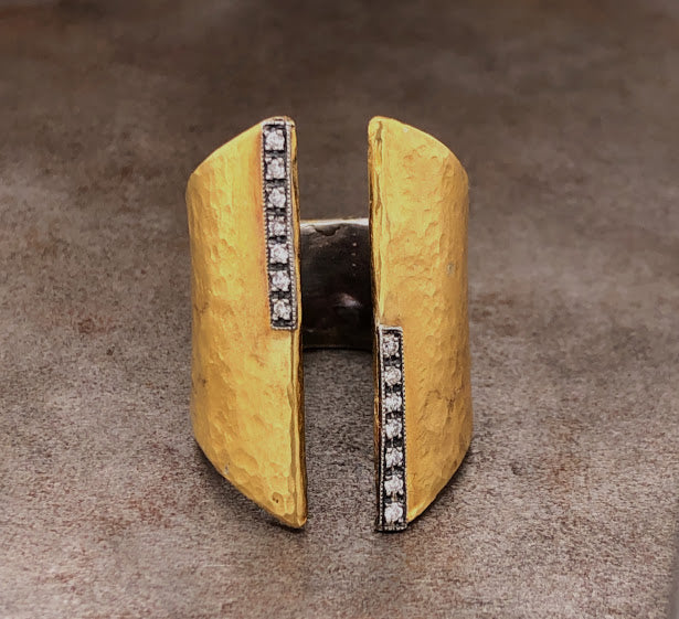 Frontal view of 24 karat yellow gold open ring. This negative space ring features an opening at the front of the ring. The ring itself spans from knuckle to knuckle and has a gradual shrinkage in the back. The ring is made from gold and has a layer of silver on the inside. On the edge of the opening that spans only half of the ring there are seven round diamonds set in oxidized sterling silver. Opposing that side has the same layout but only on the opposite side.