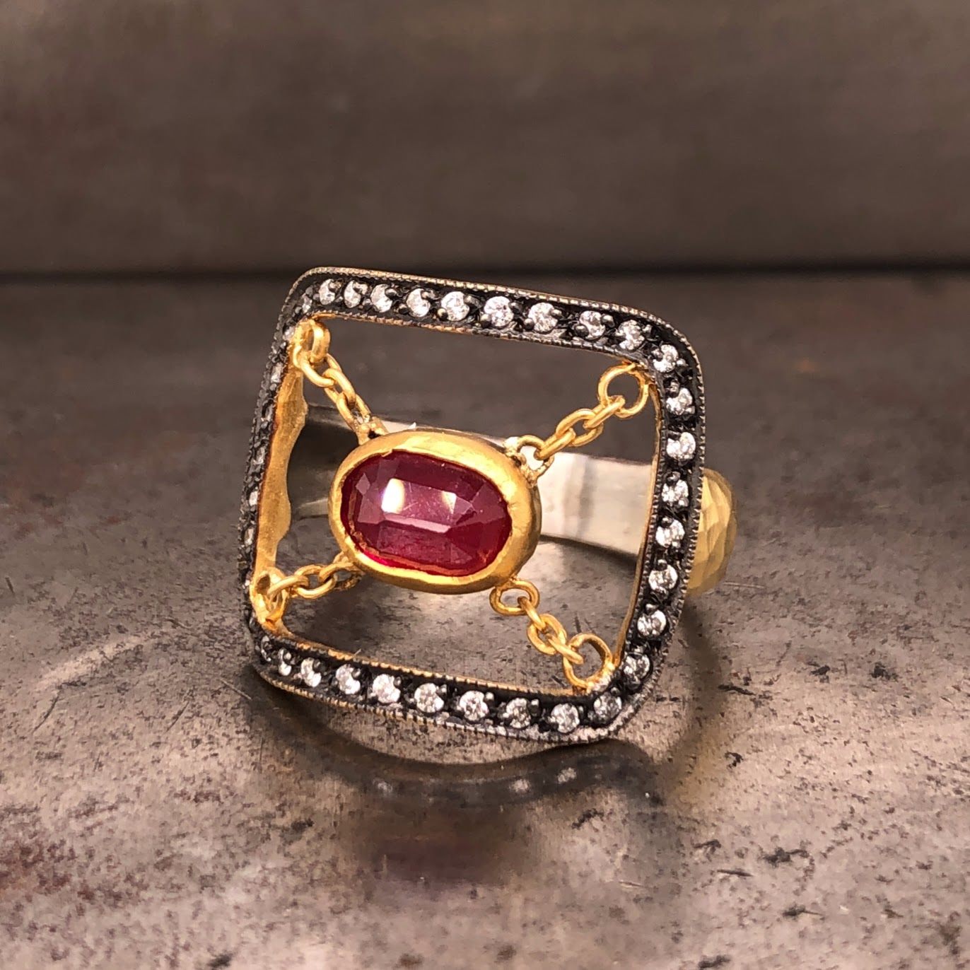 Angled view of ring laying down. There is an outline of a curved rectangle made from oxidized sterling silver that has a row of round diamonds set in it. On the inside of the rectangle, chain stretches from each corner to connect to the oval ruby that is bezel set in the center. This head lays on a gold and silver shank.