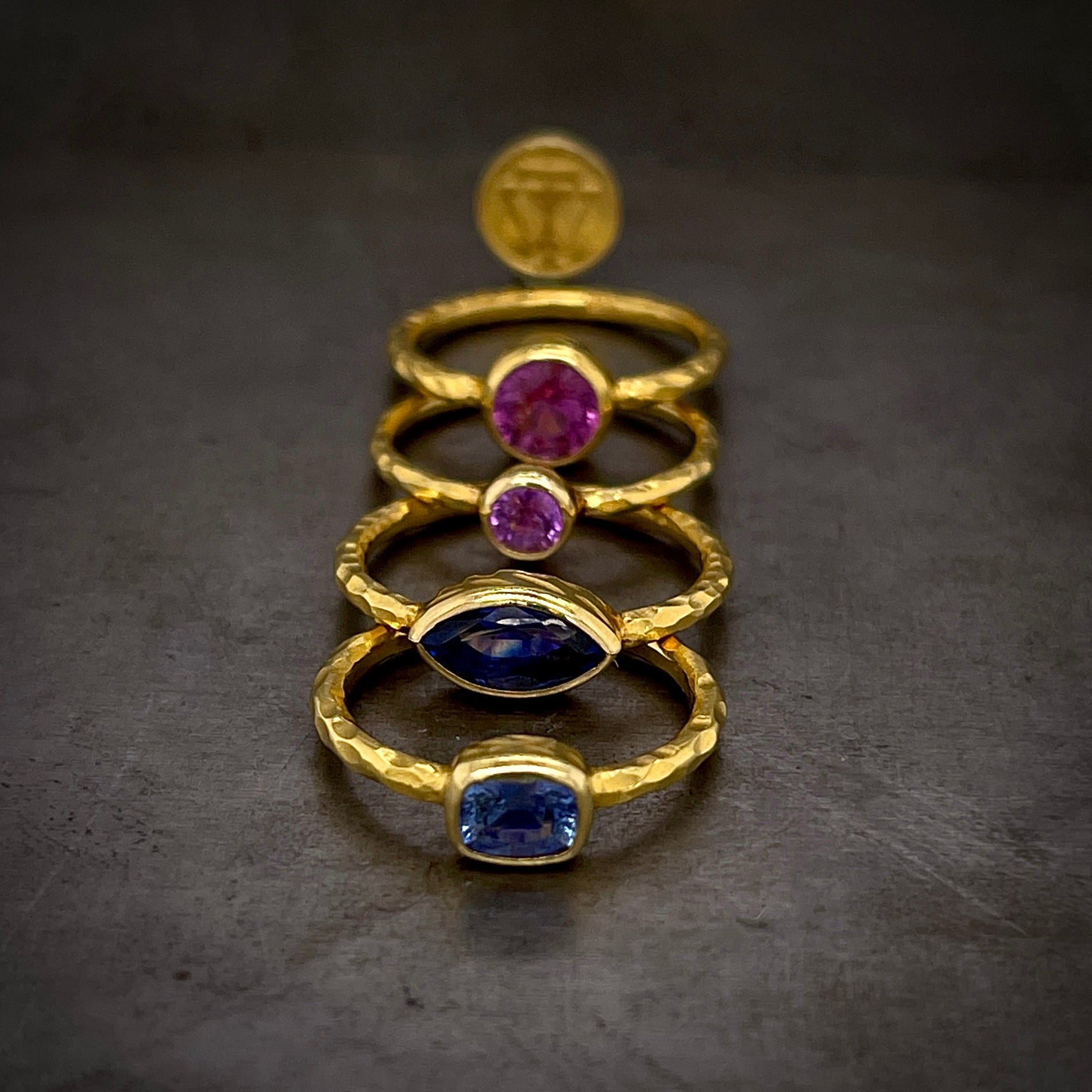 Four sapphire rings lay is a straight line with a WT gold coin in the background. The top right is the larger round pink sapphire, then the smaller lighter pink sapphire, the larger darker marquise and then the emerald cut light blue sapphire.