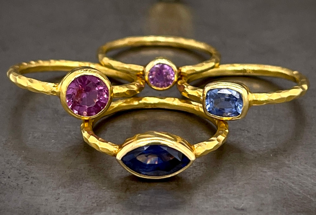 Group of four hammered sapphire rings are played out in a diamond form, laying on top of one another. The bottom ring has a deep blue marquise cut sapphire, bezel set in gold with a hammered shank. The left most ring features a round deep pink sapphire. The right ring features an emerald cut light blue sapphire and the top ring features a smaller light pink sapphire. All the stones are bezel set and feature a hammered shank.