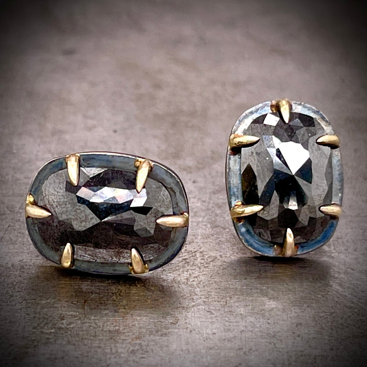 Frontal view of black diamond rose cut earrings, the left laying on its side and the right standing straight. They each feature one oval rose cut black diamond. the oxidized sterling silver expands beyond the diamonds and then features six gold prongs that encompass the diamonds.