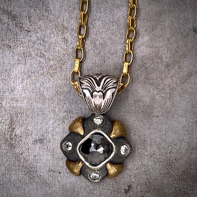 Close up view of Black Diamond Gothic Pendant. A diamond made from oxidized sterling silver is the base of the pendant. In the center is a diamond shaped black diamond. At each corner is one round brilliant diamond. On each side a claw of gold encompasses the silver. An etched bail is attached to the pendant. Lays on a chunky gold curb chain.