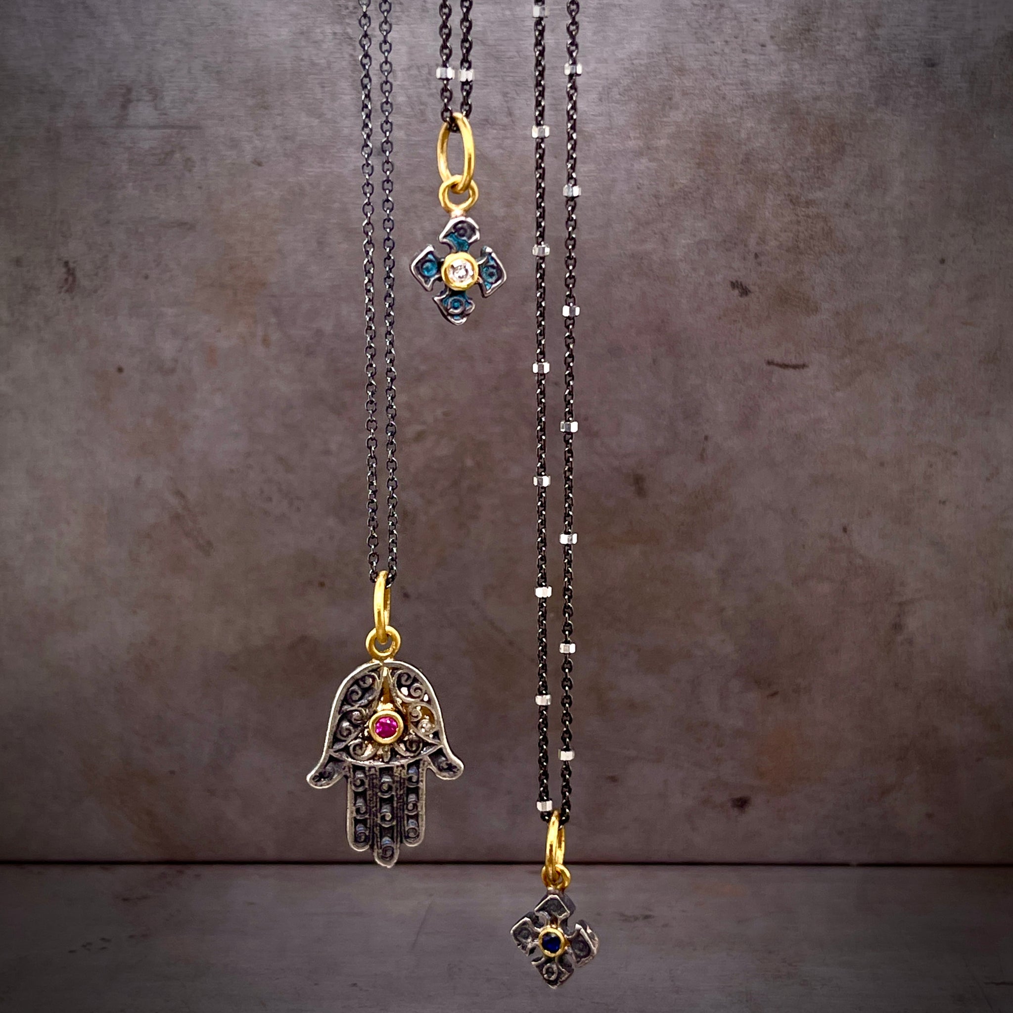 Three necklaces hang at different lengths in the air. The left is a hams hand made from oxidized silver with a round ruby set in yellow gold in the center. The center necklace is a cross with a blue coating over its silver and a round diamond set in its center. The right pendant is a crosse with a round blue sapphire set in yellow gold.