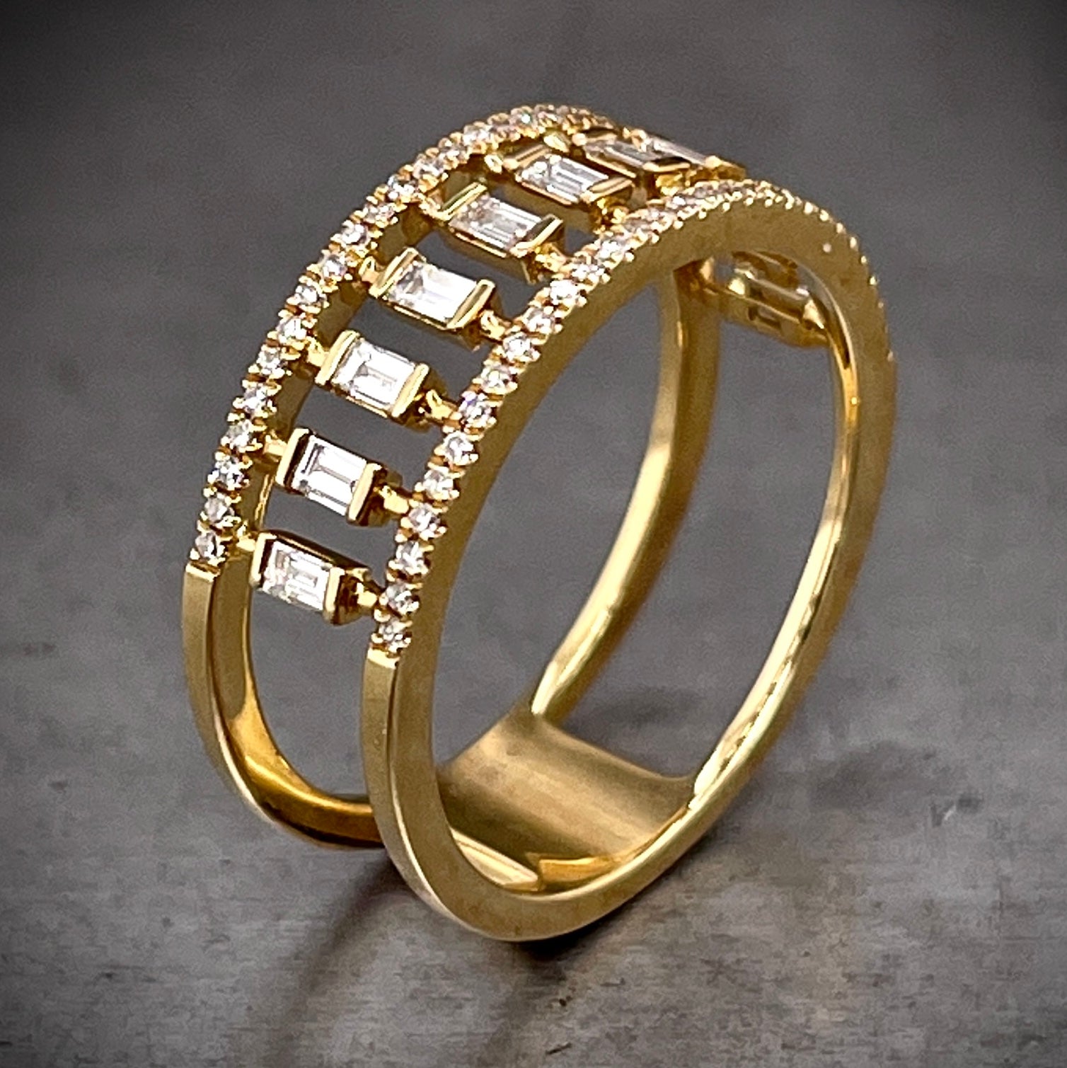 Angled aerial view of Baguette Diamond Cigarette Band. Here you can see baguette diamonds partial bezel set in the middle of the band. These baguettes travel across the shoulders of the ring but do not continue onto the back. Opposing either side of the baguettes is a row of round brilliant diamonds prong set. There are two shanks that form from the round diamonds and connect in the back with a block of gold.