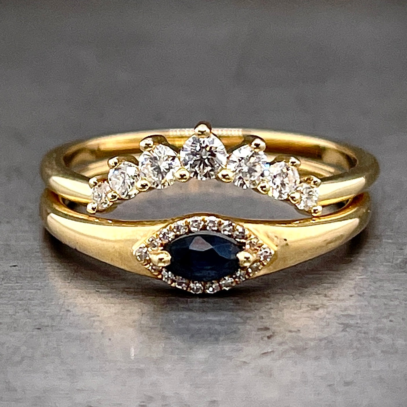 Two rings lay on top of one another. The marquise sapphire ring lays on the bottom with a diamond tiara ring laying on top of it. The diamond tiara ring features seven round brilliant diamond three prong set and create the curve of a tiara.