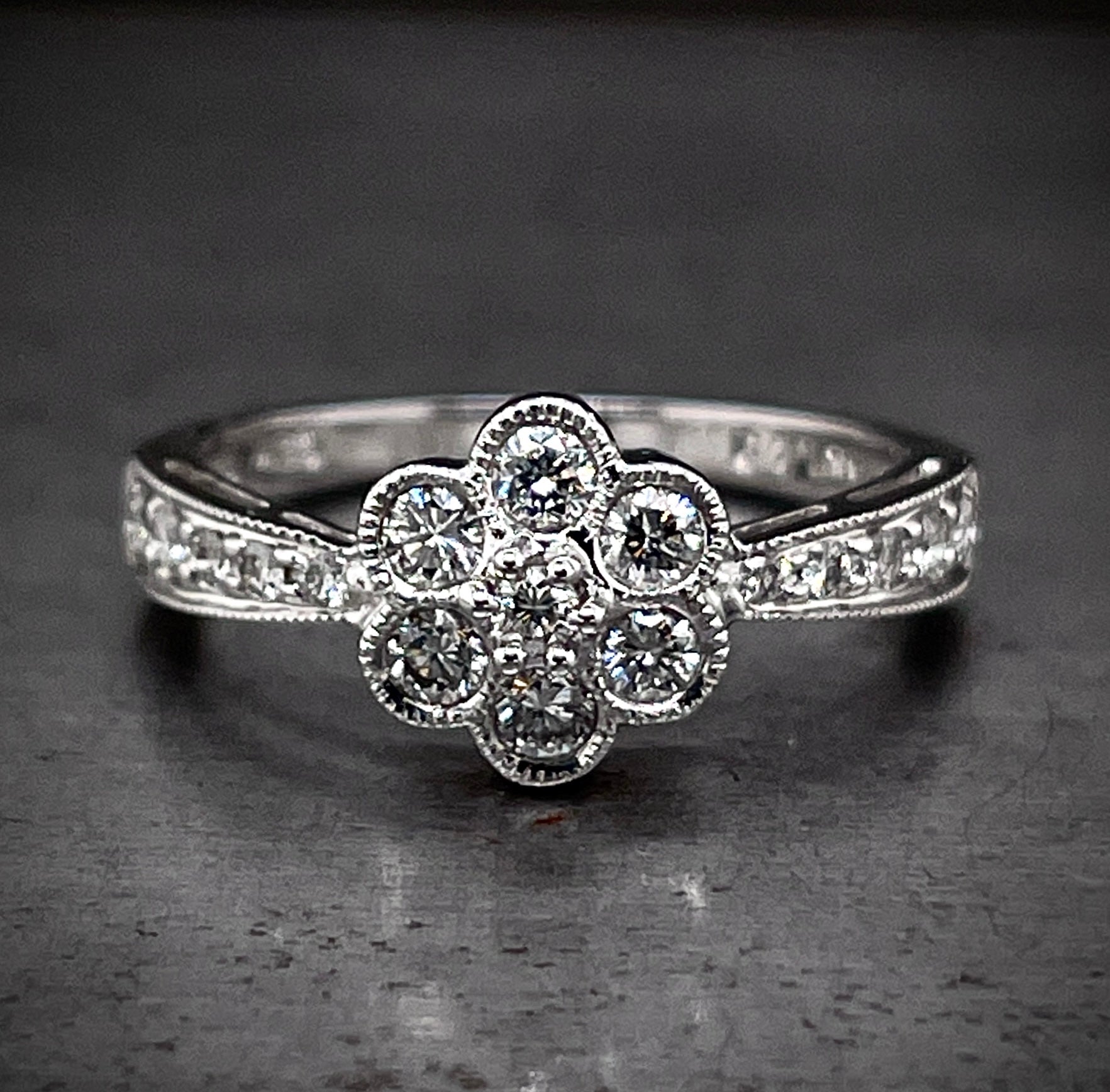 Full view of diamond flower ring. The center features seven round brilliant diamonds that create the shape of a flower (one in the center and six that surround it). Flanking either side of the flower the band expands from a smaller width to a larger width, diamonds are set in the band and increase in size with the band.