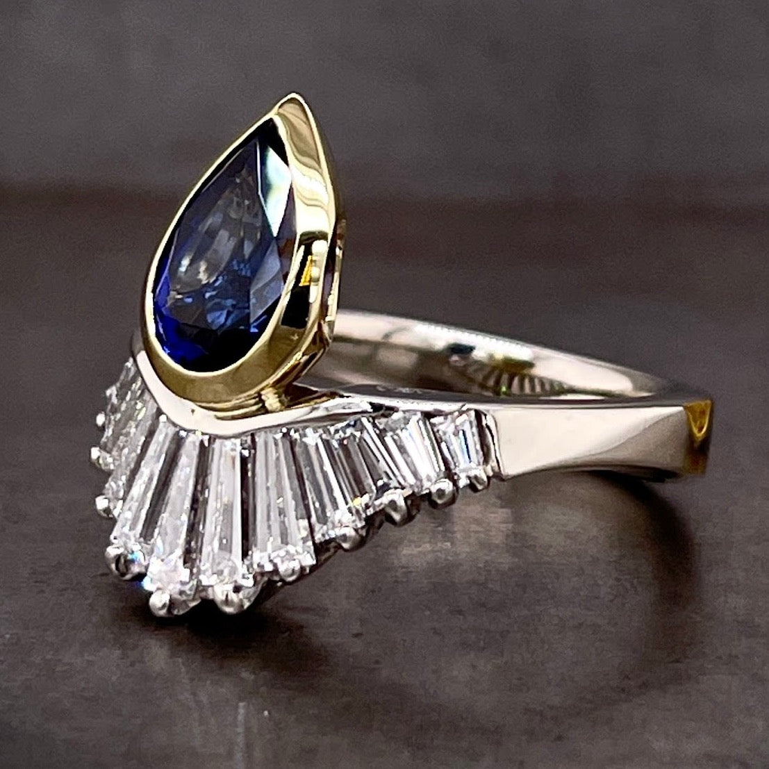 3/4 view of blue sapphire and diamond ring. The blue sapphire is positioned upwards.  The tapered diamond baguettes are  arrayed along the lower half of the ring.  
