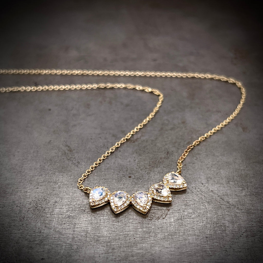 Partial view of diamond and moonstone necklace. Pendant features five pear cut cabochon moonstones. Each moonstone has a halo of diamonds surrounding it. The five stone create a slight curve, creating an obtuse U shape. The chain is attached to either side of the pendant and snakes through the image.