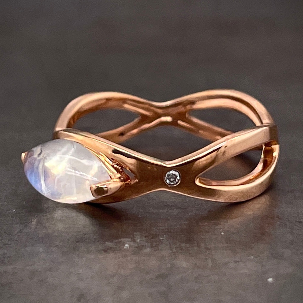 3/4 profile view of moonstone and diamond ring. This ring features an open woven shank made from 14 karat rose gold. The top of the ring features a marquise cut cabochon moonstone, two prong set on its western and eastern girdles. The moonstone is positions western to eastern. On the shoulder of the ring, there is a round brilliant diamond that is flush set into the shank.
